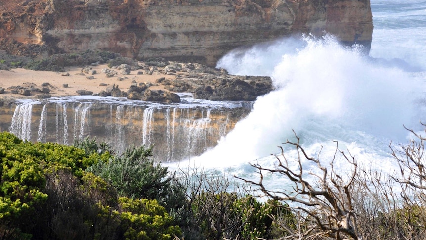 Large waves crash over seas cliffs and cascade into the water in a series of waterfalls.