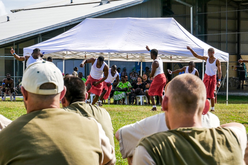 A group of men wearing khaki t-shirts sitting on the ground watching Indigenous performers.