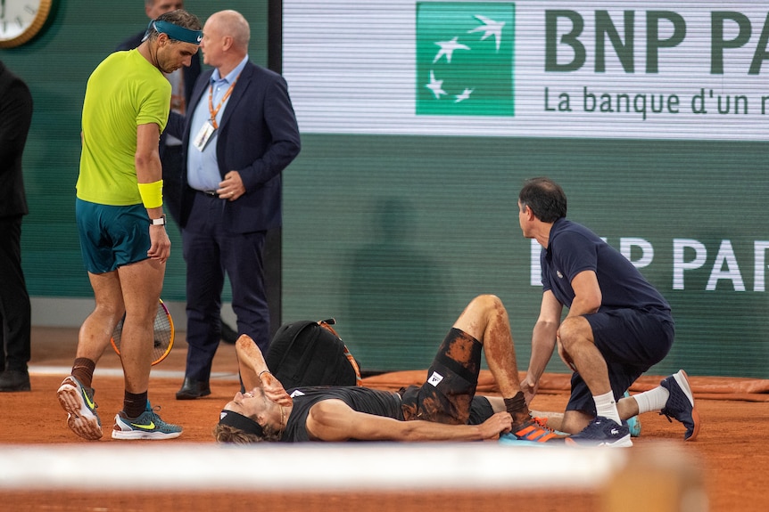A male tennis player wearing green looks down at his opponent wearing black and clasping his foot