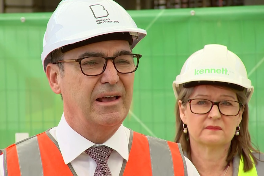 A man with glasses wearing a hard hat in front of a green background