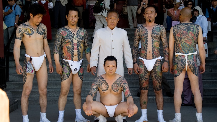 Yakuza members with white loincloths show off their intricate tattoos in public. Their boss stands behind them in a white suit.