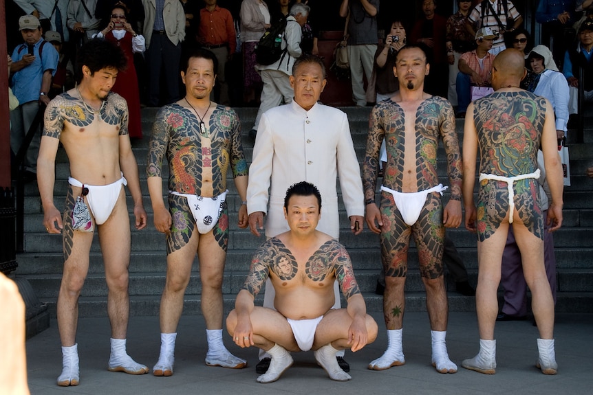 Yakuza members with white loincloths show off their intricate tattoos in public. Their boss stands behind them in a white suit.