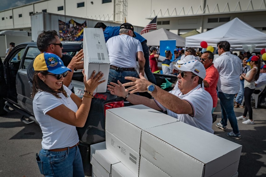 A woman wearing a cap and sunglasses passes a box to a man who is stacking boxes, with people unloading supplies in background