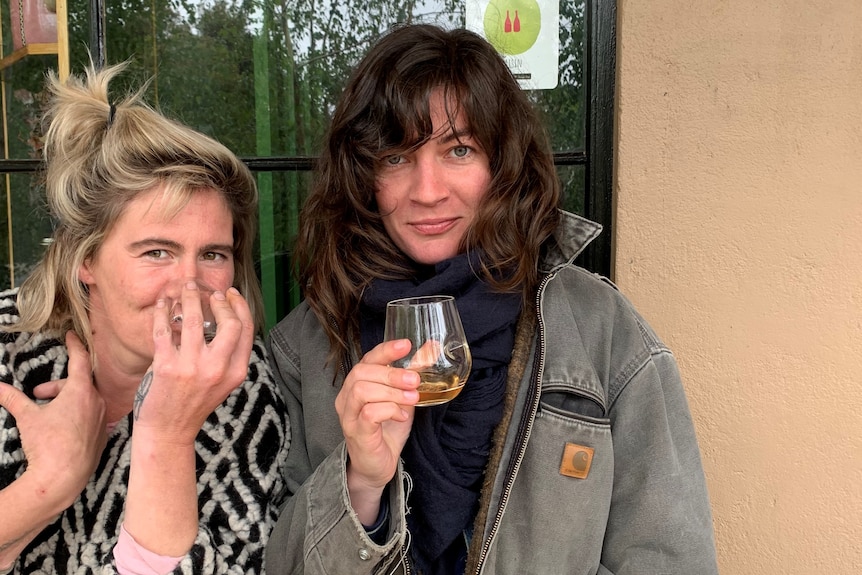 A blonde woman holds a glass to her face next to a brunette woman who holds a wine glass, both sitting in front of a window