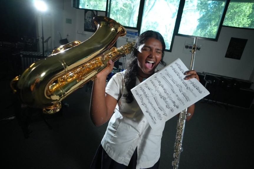 Girl in school uniform holds up a saxophone in one hand, flute and sheet music in the other, while appearing to sing.