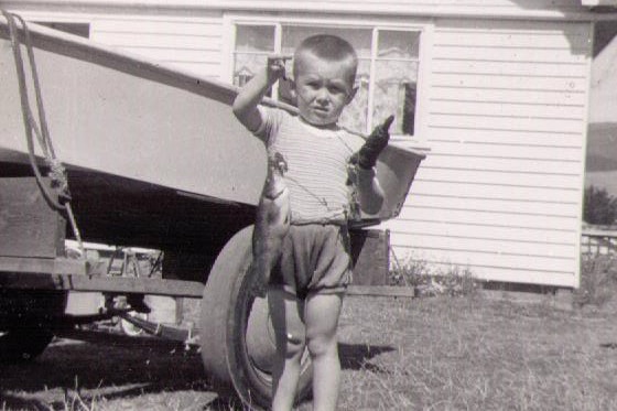 A young boy stands in front of his house holding a fish in a black and white photo