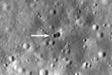 A photo of the Moon, where a small white arrow points to a double crater.