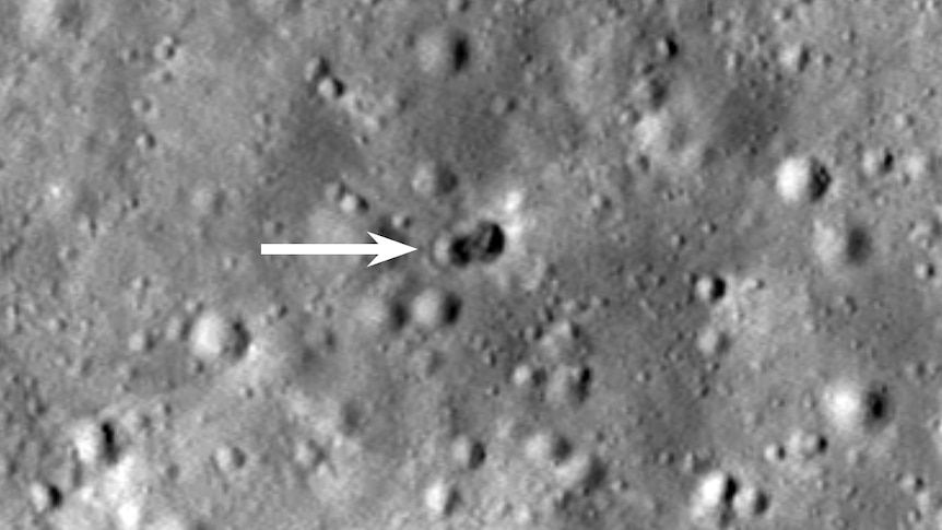 A photo of the Moon, where a small white arrow points to a double crater.