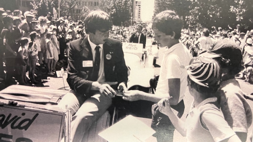 A black and white photo of a man in a jacket and tie on a parade float giving his autograph to some children.