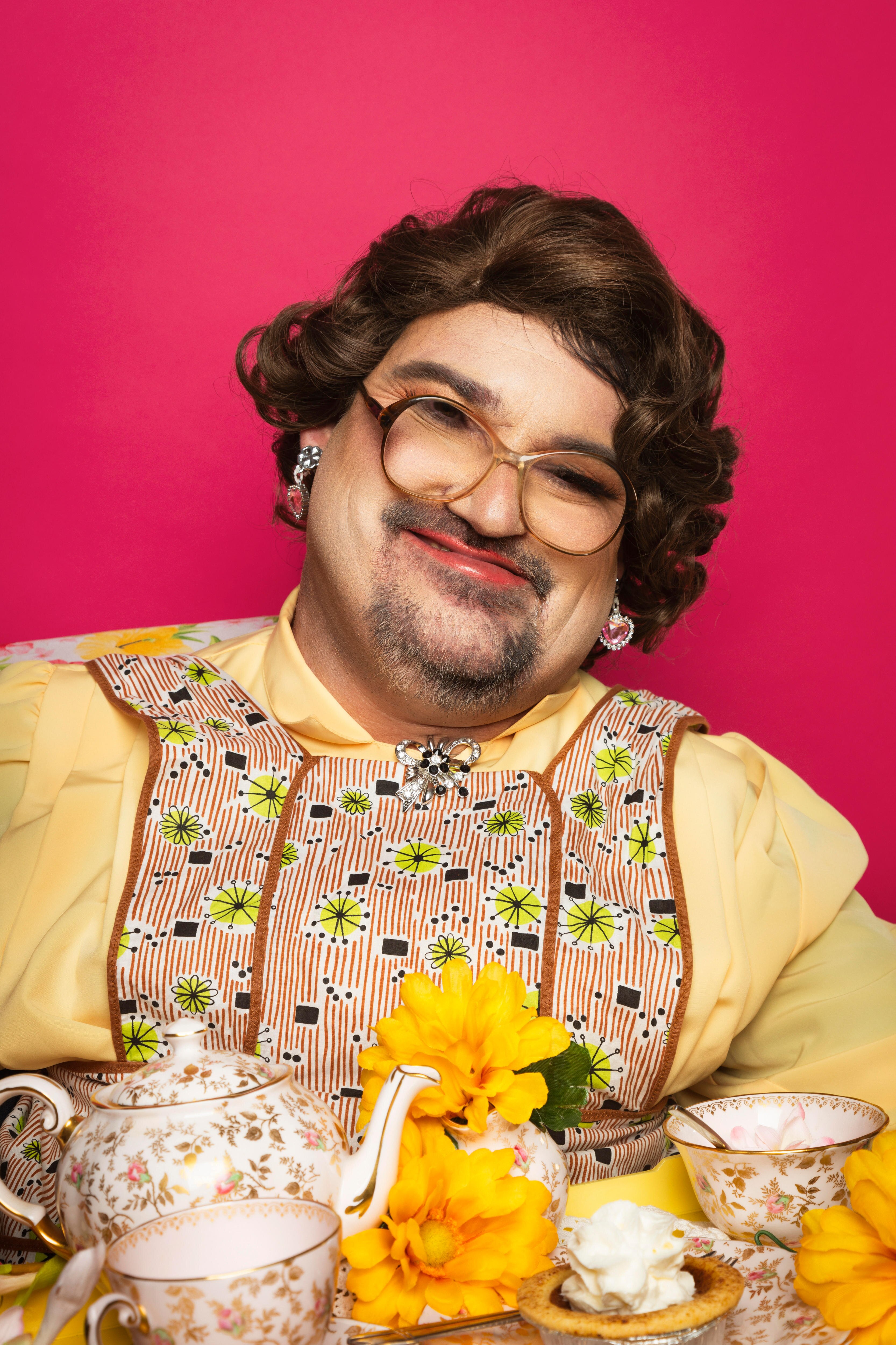 A man, dressed as a woman with glasses, a yellow top and apron, holding a tea set.