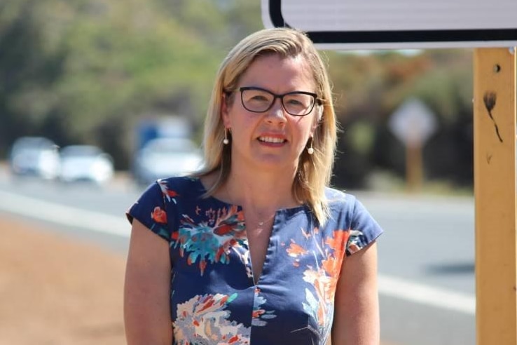 A blonde woman with glasses stands on the side of a road