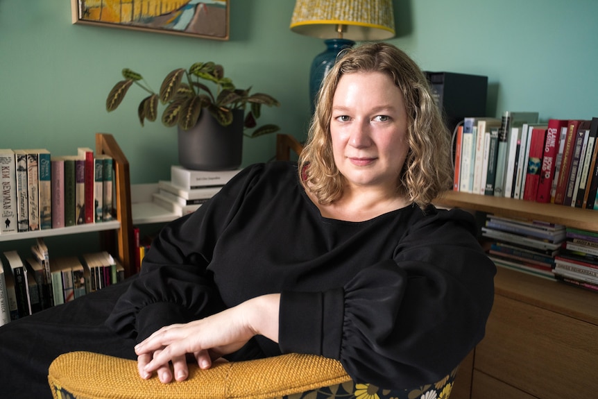 a blonde woman wearing all black sitting at a desk with bookshelves filled with books and plants behind her