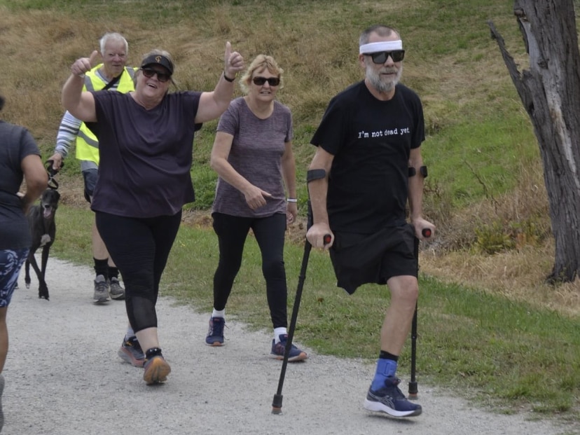 A man with one leg is using crutches to compete in parkrun. A woman behind him points two thumbs up to the camera.