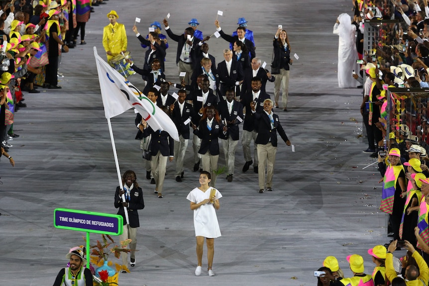 An athlete carrying a flag of the Olympic rings leading a team of athletes at an opening ceremony for the Olympics. 
