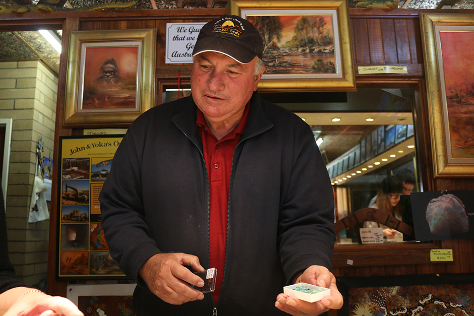 A man stands behind a store counter, holding a small box of opal gemstones.