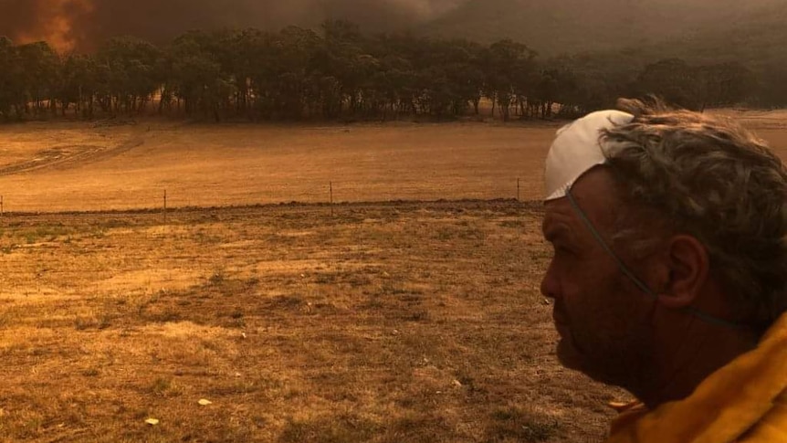 A firefighter with his mask pulled up to his forehead looks on at a raging fire on the horizon.