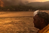A firefighter with his mask pulled up to his forehead looks on at a raging fire on the horizon.