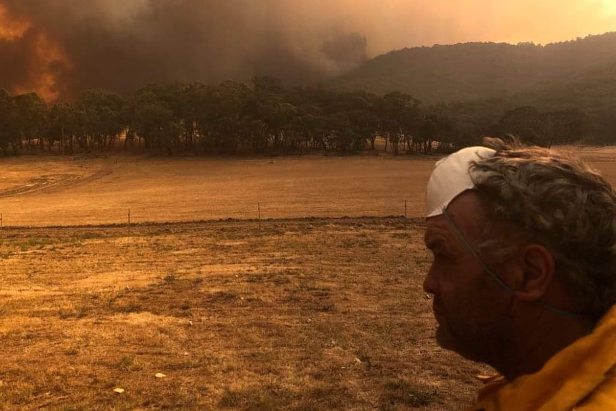 A firefighter with his mask pulled up to his forehead looks on at a raging fire on the horizon.