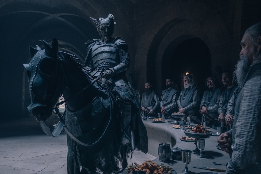 A mythical creature resembling a man but with a wooden face wears knight's armor and addresses medieval round table atop a horse