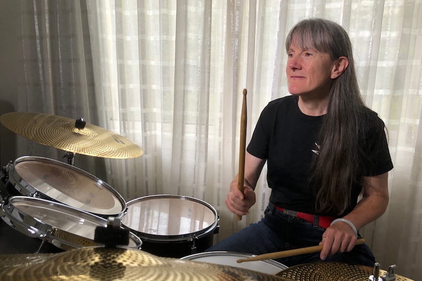 A woman with grey hair and a fringe sits at a drum set