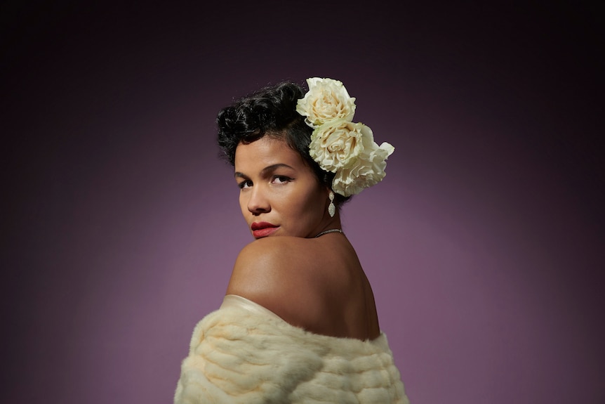 A Jamaican Australian woman looks over her shoulder into the camera, with white flowers in her carefully pinned-back hair