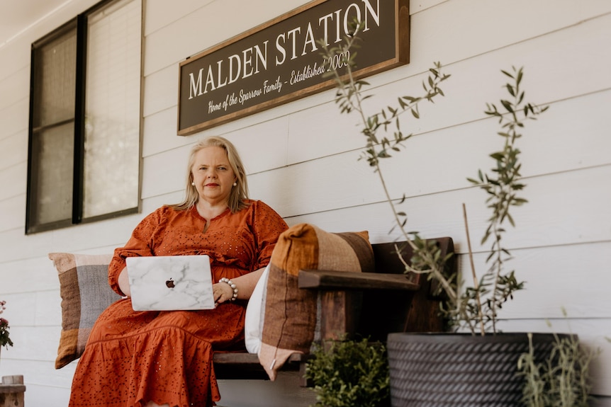 A lady in a rust coloured dress sits with her laptop on the verandah.