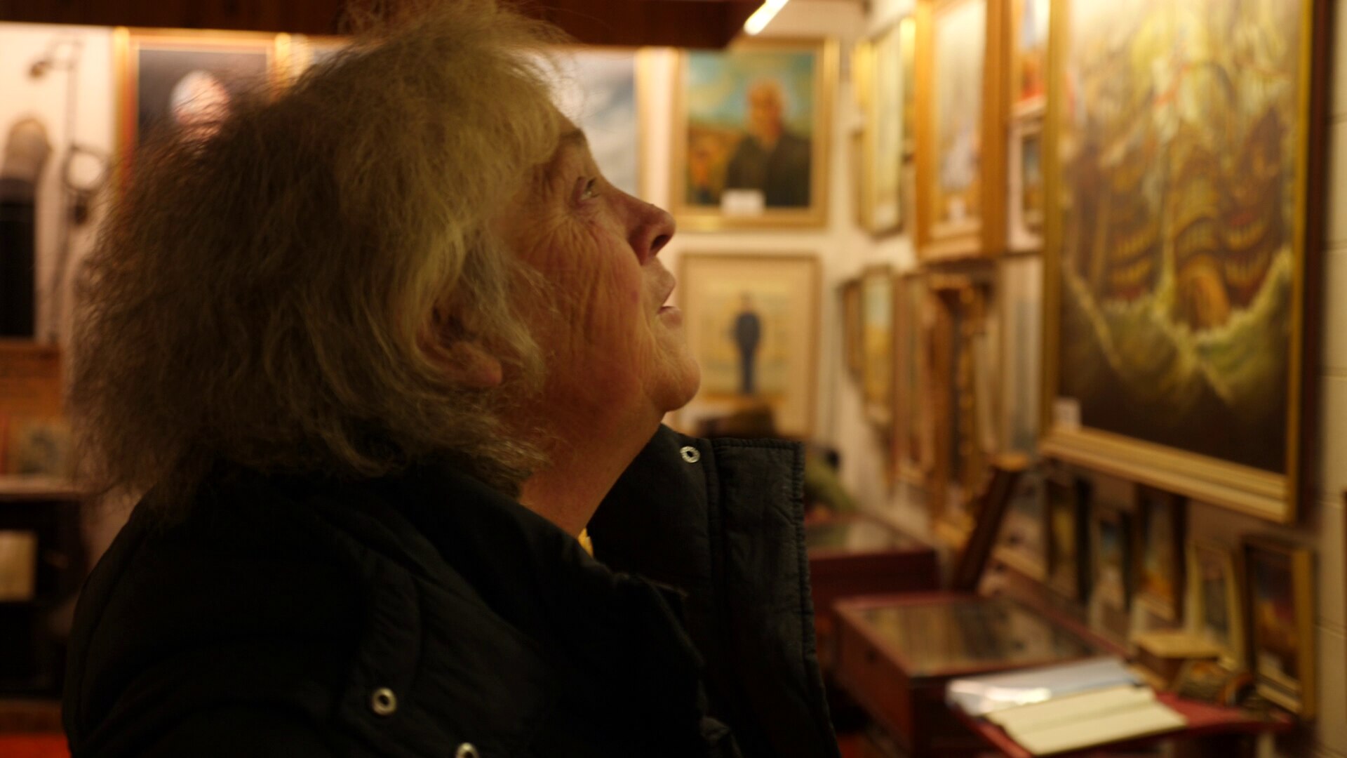 An older woman in a dark coat gazes at paintings on the wall of an art gallery.