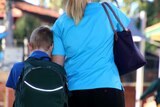 A mother in a blue top walks with her son carrying a green backpack to drop him off at school.