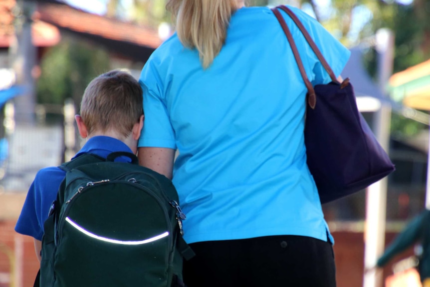 A mother in a blue top walks with her son carrying a green backpack to drop him off at school.