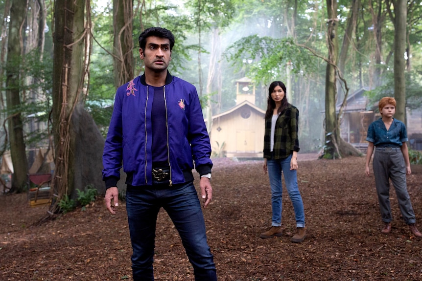 Kumail Nanjiani, Gemma Chan and Lia McHugh are standing looking alert at a seemingly abandoned campsite in the woods