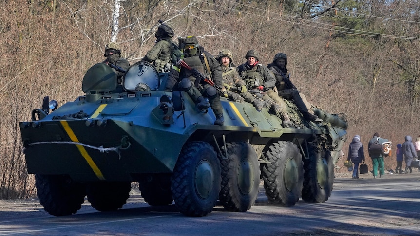 A group of Ukrainian soldiers with yellow armbands ride through a street on top of an armoured vehicle.