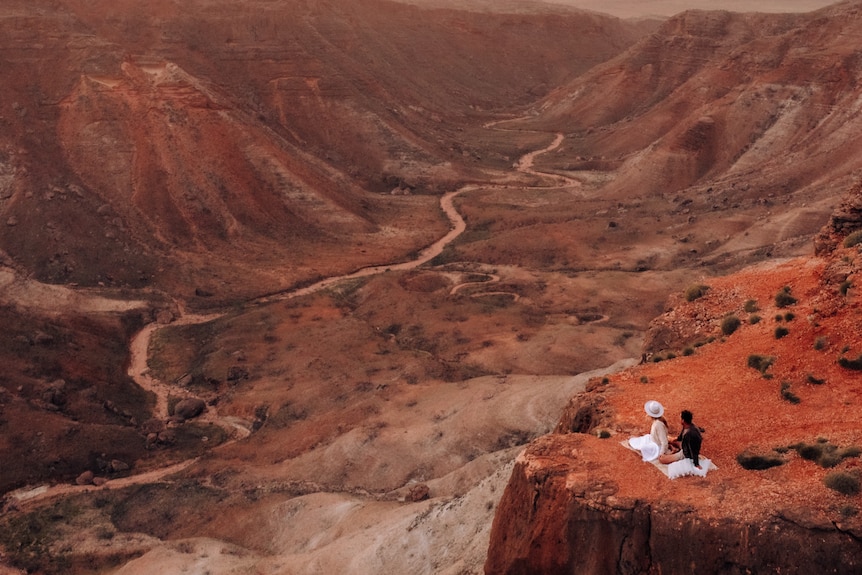 Two people sit on a picnic rug overlooking a canton with a road passing through it. 