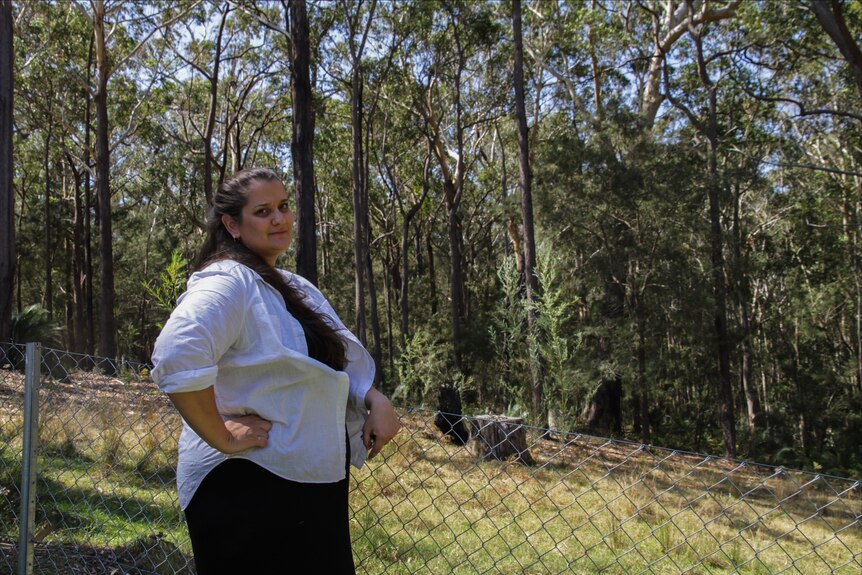 A woman in a white shirt leans against a wire fence. In the background is eucalyptus gum bushland.