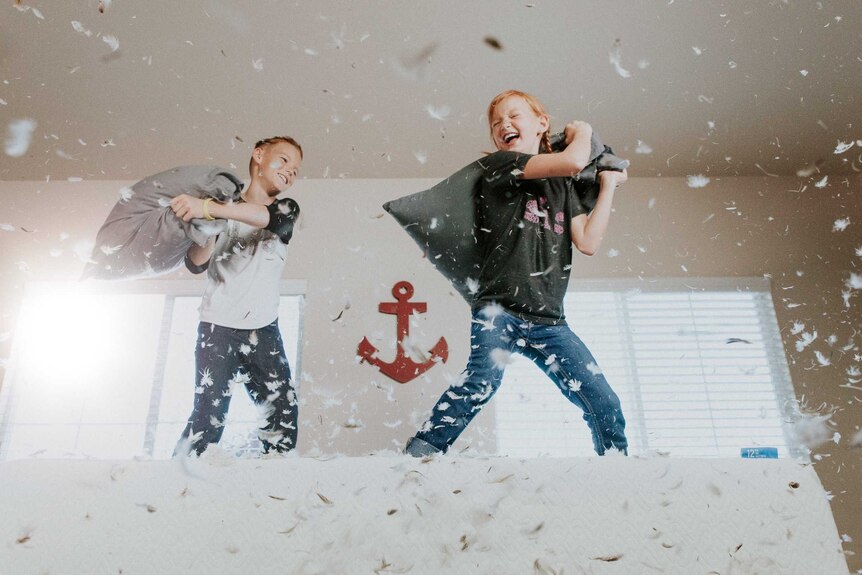 Two laughing kids having a pillow fight on top of a bed, with feathers in the air.