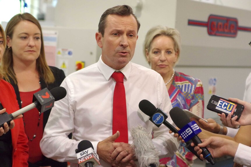 Mark McGowan stands in front of reporters holding microphones with people behind him.