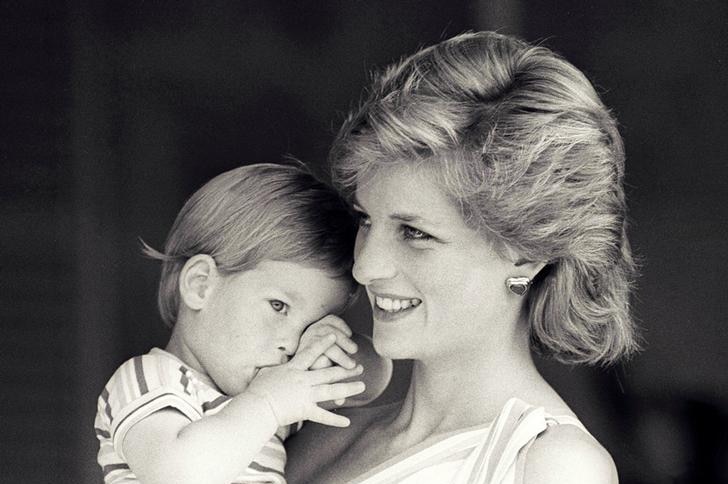 Princess Diana smiles in mid-shot as Prince Harry is in her arms while sucking on his thumb.