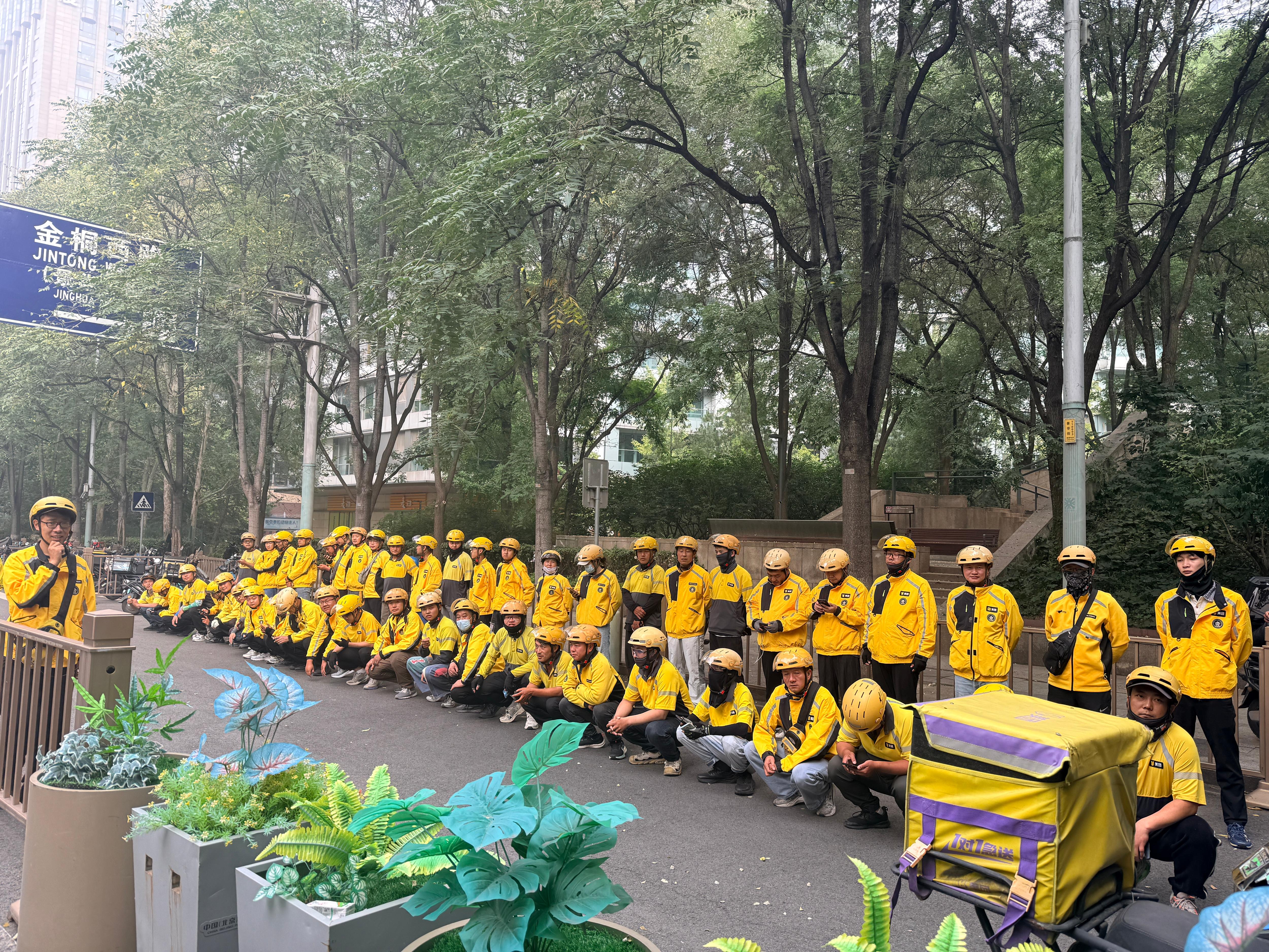 A long line of food delivery drivers in bright yellow coats stand together next to their bikes.