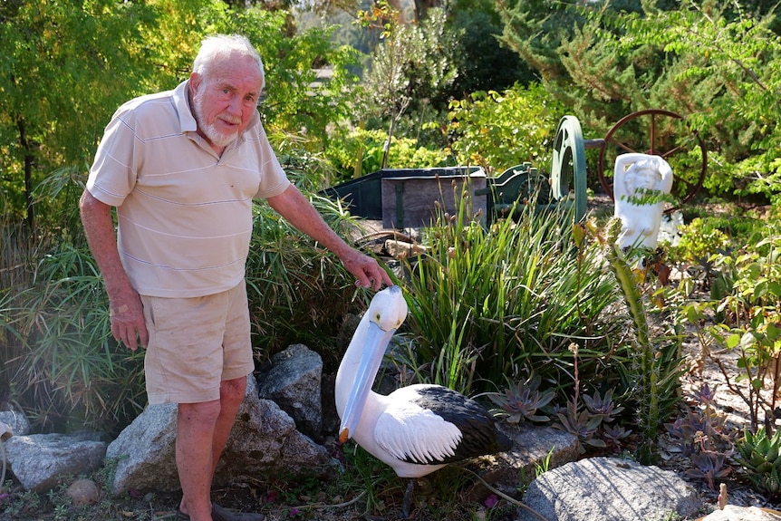 An older man stands in a garden in front of a pelican sculpture and sculpture of a nude woman.