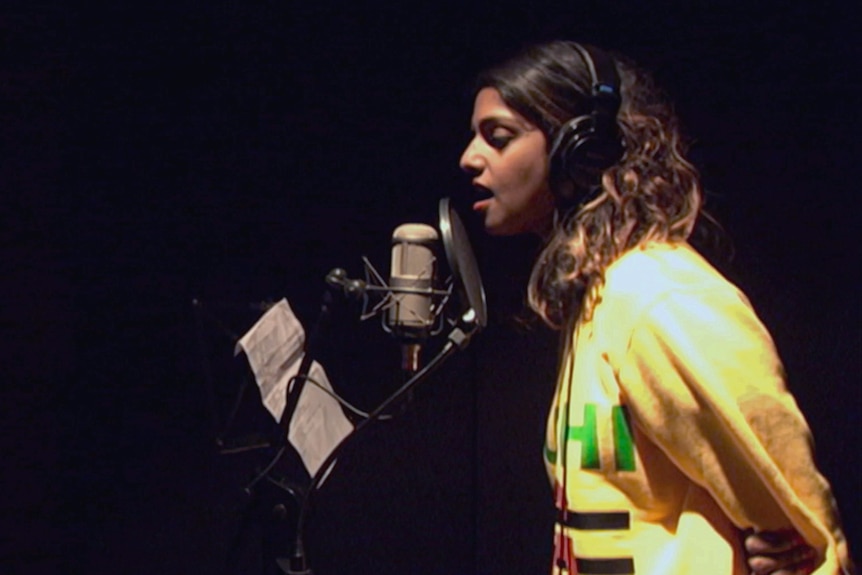 Singer M.I.A. with hair out and wearing yellow graphic sweater, singing into mic in studio, eyes closed.