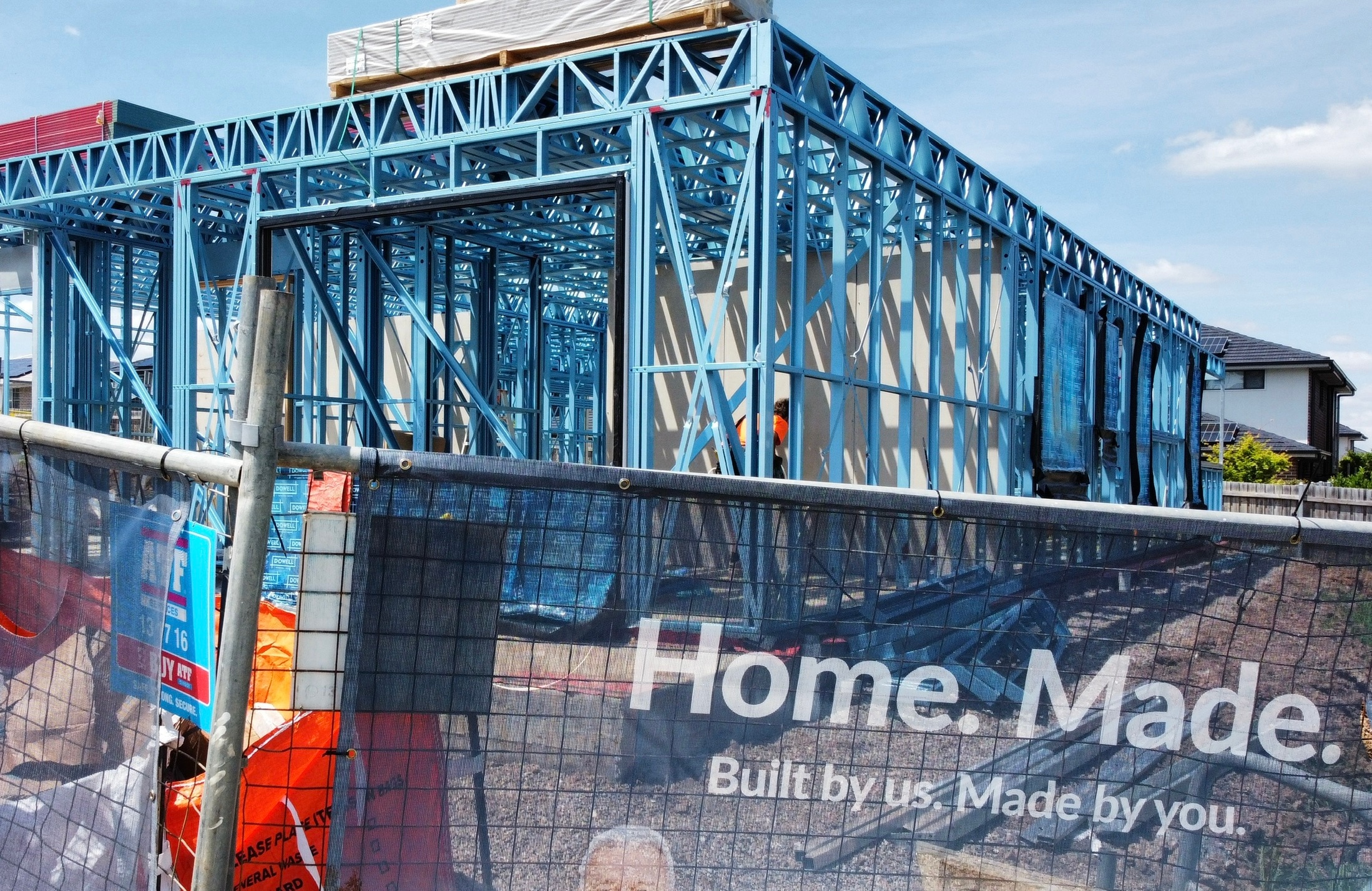 A steel frame of a house, a iron mesh fence around it with Home. Made. Built by us. Made by you. logo.