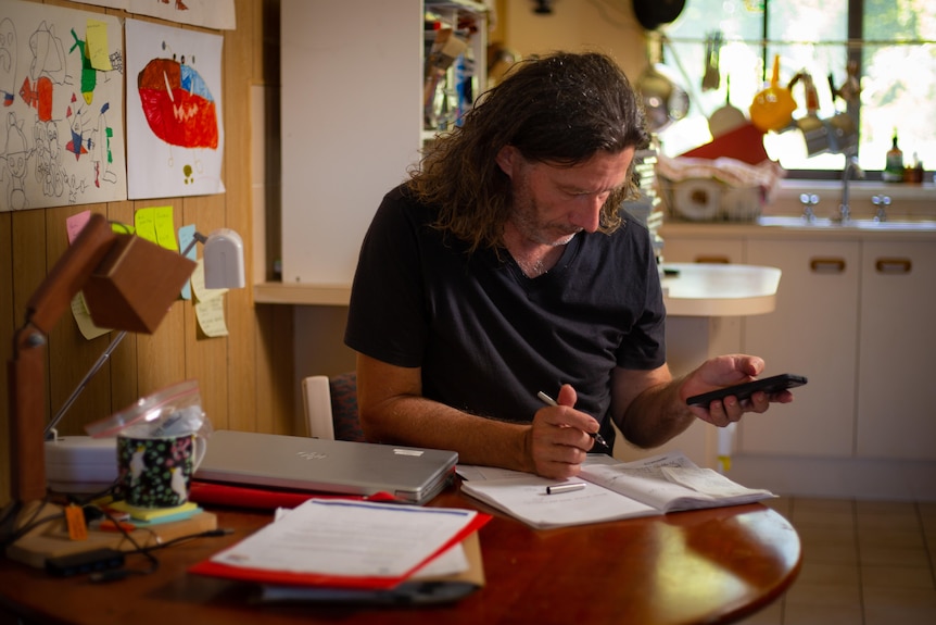 A middle-aged white man with long hair sitting at a table, surrounded by papers