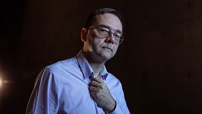 Man wearing glasses and button up shirt with left hand clutching chest, standing in darkly lit theatre space.