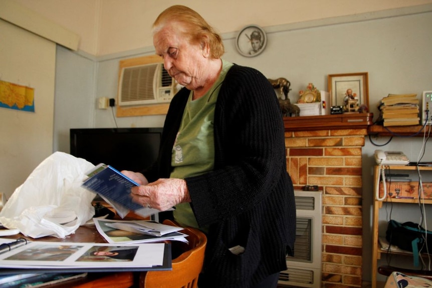 An older woman wearing a black cardigan goes through paperwork at a table. A radiator heater glows red in the background.