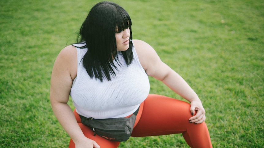 A woman with black hair is kneeling on the grass and stretching her legs. She is wearing exercise clothing.