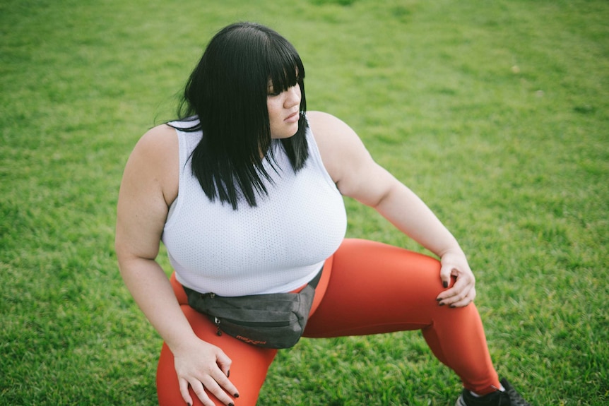 A woman with black hair is kneeling on the grass and stretching her legs. She is wearing exercise clothing.