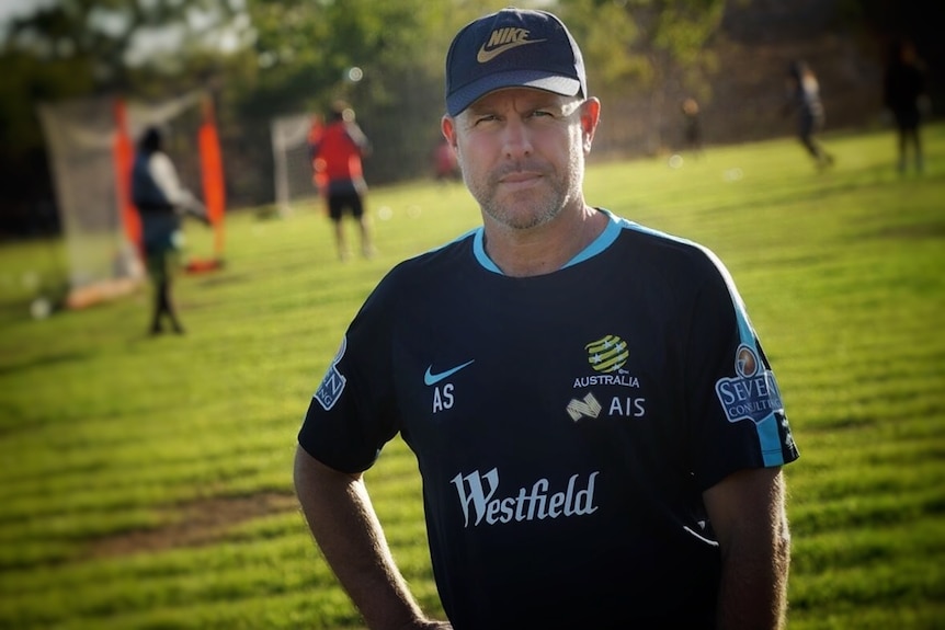 Alen Stajcic wears sports clothes and a Nike cap and looks into the camera, while standing on a soccer field