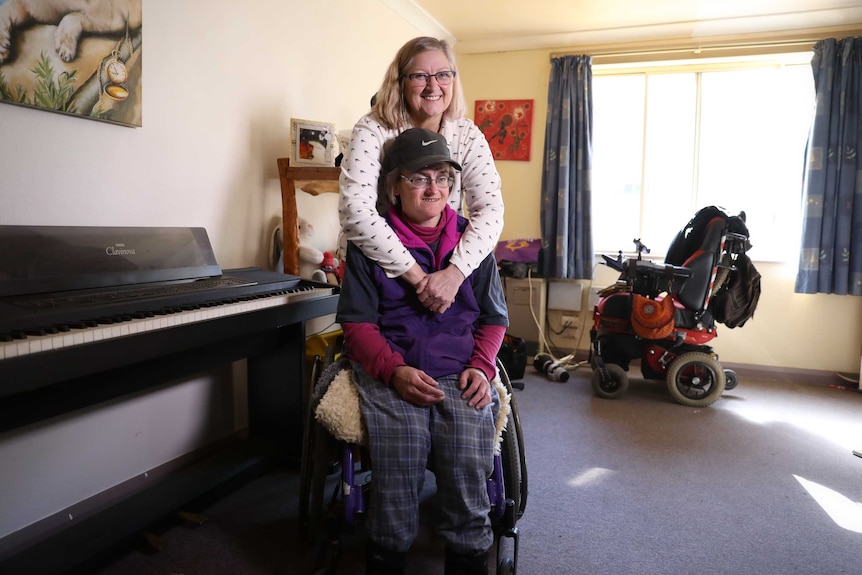 A smiling mother hugs her adult daughter, who is in a wheelchair.