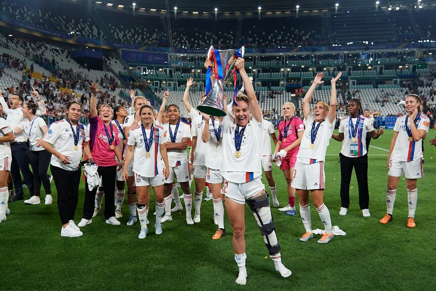 Ellie Carpenter, with a brace on her left leg, lifts the Champions League trophy in front of her Lyon teammates.