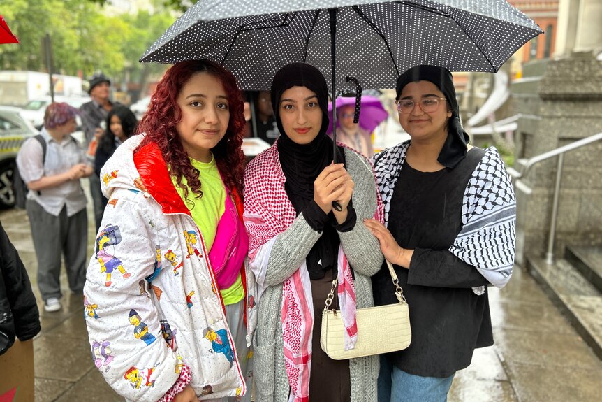 Three women holding an umbrella smile.
