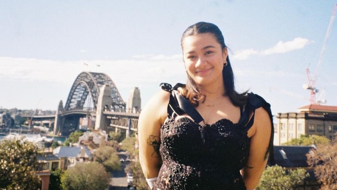 A young woman with black hair and in a black skirt and top stands on a hill with the Sydney CBD in the background. 